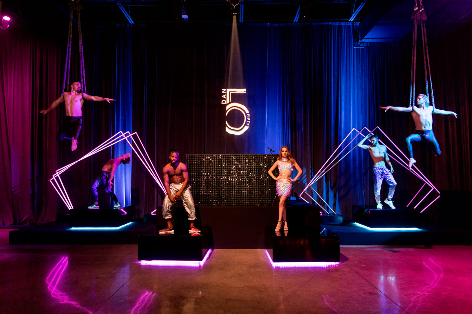 Aerial and dance cast performing on a neon-framed stage at a 50th birthday celebration